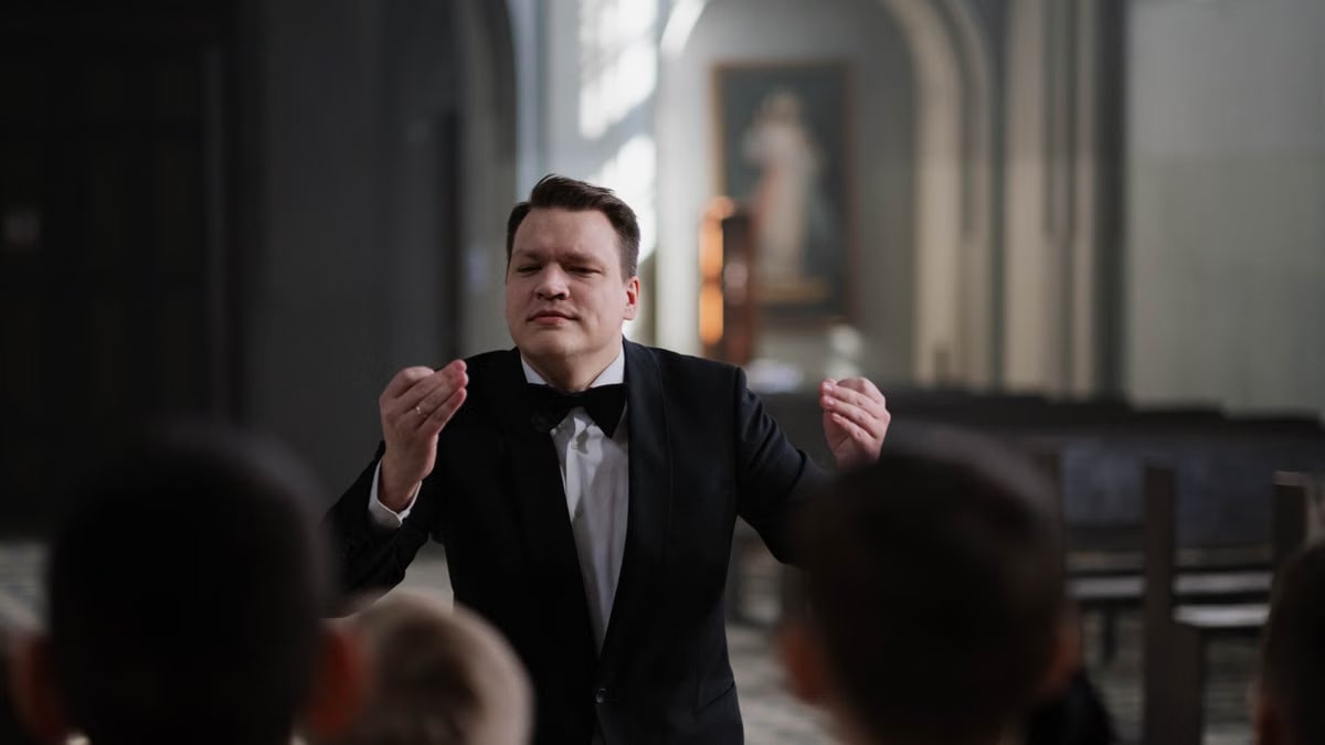 A male conductor leads a boys' choir rehearsal in a serene church setting.
